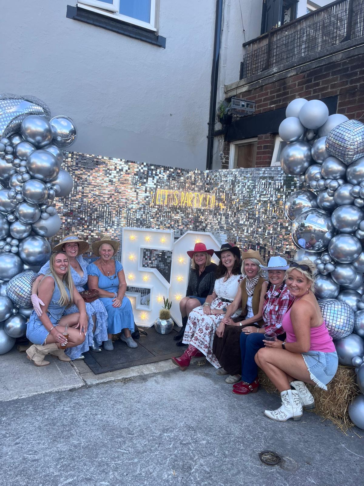 A group of women wearing cow boys hats are pictured in front of a free-standing lit up '50'. Either side of the sign and the women are silver balloon arches as well as a glitter backdrop. Sue is pictured to the immediate left of the '50' sign. 