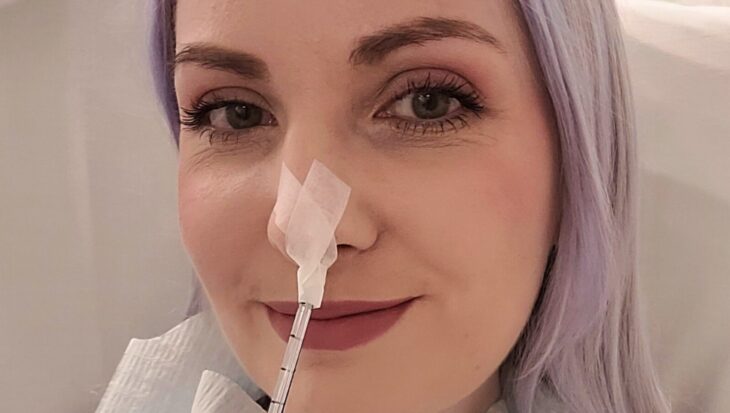 Leanne smiles at the camera in a selfie style photo. She has a feeding tube inserted into her nose and blue roll tucked into the collar of her t-shirt.