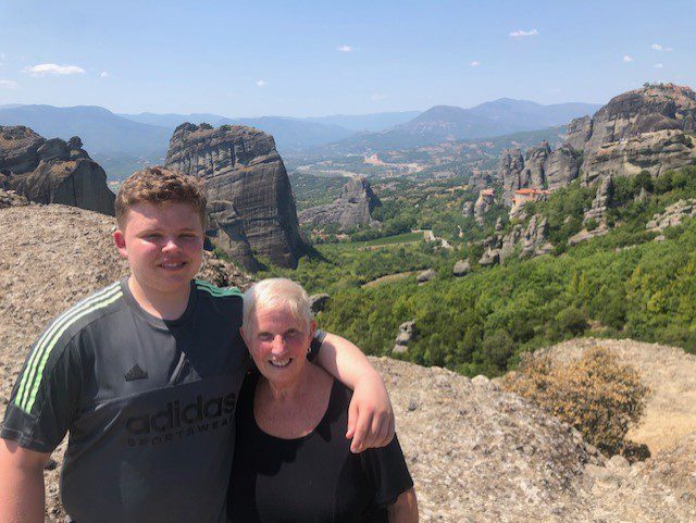 Sue (right) is pictured next to her grandson, with his arm around the top of her shoulders, they are both smiling at the camera. In the background is a hill landscape in Meteora. 