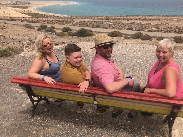 Sue (far right) is sat on a wooden bench with her adult son, teenage grandson and adult daughter sat beside her. Everyone has turned around to look at the camera and a beach and coastline are visible in the background. 