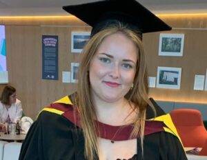 Charlotte wears a long, red patterned skirt, with a black top, and a black graduation gown and cap. Her graduation gown has yellow and red round the shoulders. She smiles, and has long, blonde straight hair.