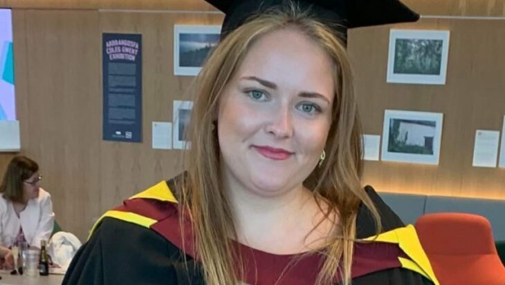 Charlotte wears a long, red patterned skirt, with a black top, and a black graduation gown and cap. Her graduation gown has yellow and red round the shoulders. She smiles, and has long, blonde straight hair.