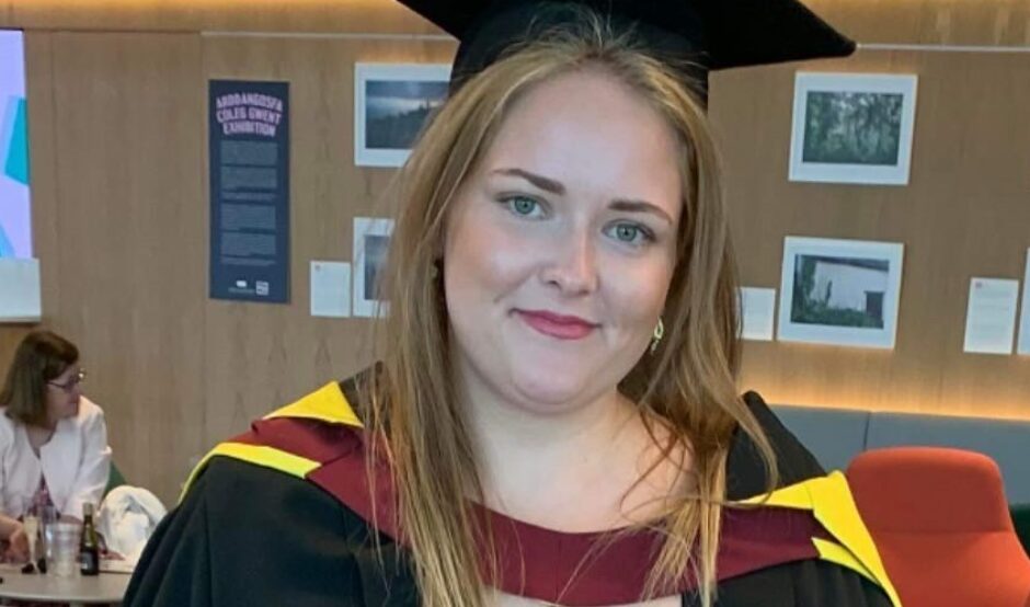 Charlotte wears a long, red patterned skirt, with a black top, and a black graduation gown and cap. Her graduation gown has yellow and red round the shoulders. She smiles, and has long, blonde straight hair.