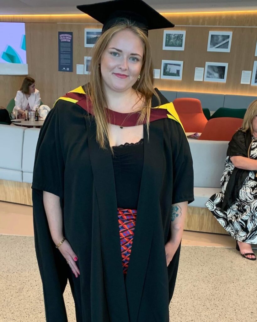 Charlotte wears a long, red patterned skirt, with a black top, and a black graduation gown and cap. Her graduation gown has yellow and red round the shoulders. She smiles, and has long, blonde straight hair.