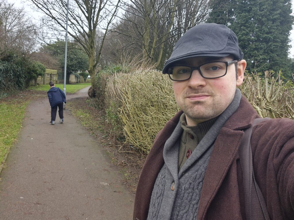 Callan is stood in a park, taking a selfie. He wears a brown jacket, with a thick, cable knot, grey cardigan underneath. He also wears a grey flat cap and black, thick rimmed glasses. He has a bit of facial hair around his mouth.