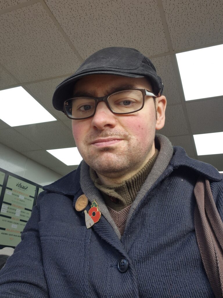 Callan is wearing a navy, thick, blazer jacket and a matching flat cap. He has a pin badge for the poppy appeal on his jacket.