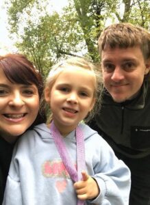 Chris, Rachel, and their daughter Ella-Rose are smiling while standing outdoors in a green, wooded area, looking happy and relaxed.