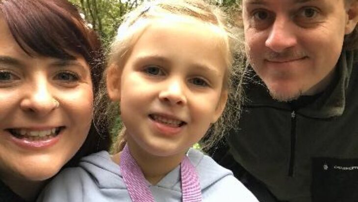 Chris, Rachel, and their daughter Ella-Rose are smiling while standing outdoors in a green, wooded area, looking happy and relaxed.