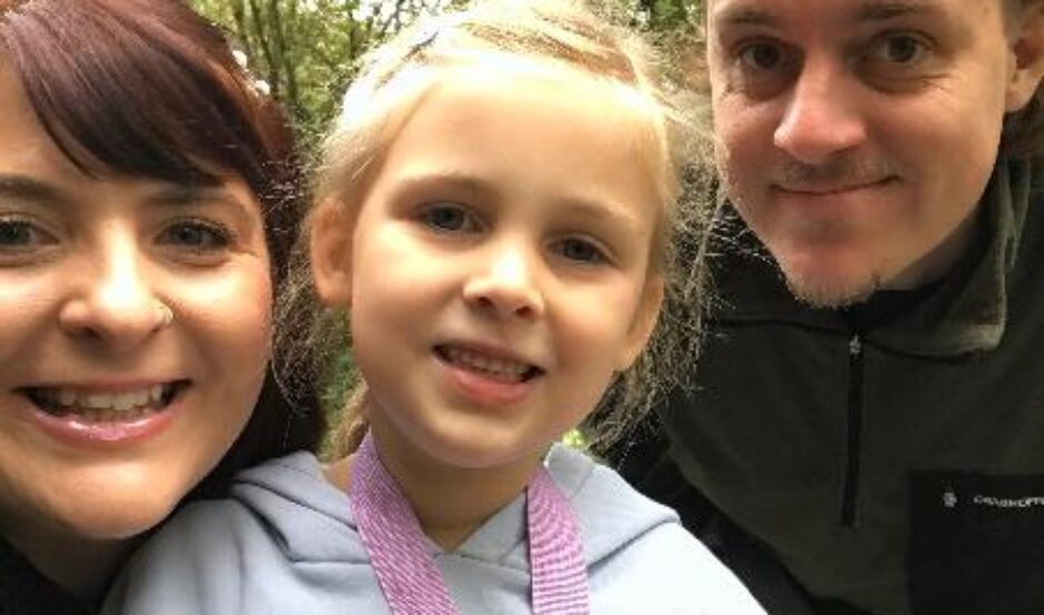 Chris, Rachel, and their daughter Ella-Rose are smiling while standing outdoors in a green, wooded area, looking happy and relaxed.