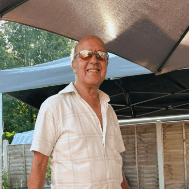 A man in a a white shirt (Charlie) smiles at the camera. Charlie is in the garden and is stood in front of a patio table, he is wearing oven gloves and holding a cooking utensil.