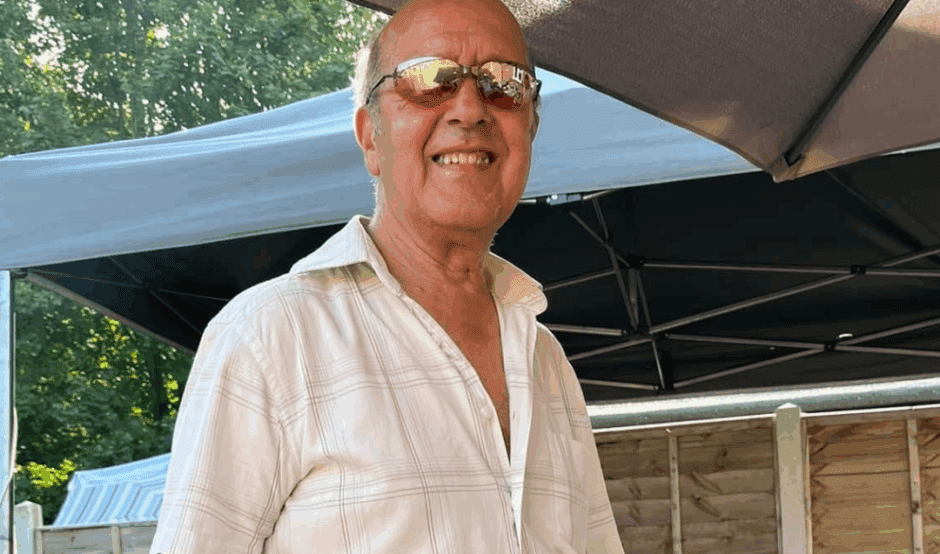 A man in a a white shirt (Charlie) smiles at the camera. Charlie is in the garden and is stood in front of a patio table, he is wearing oven gloves and holding a cooking utensil.