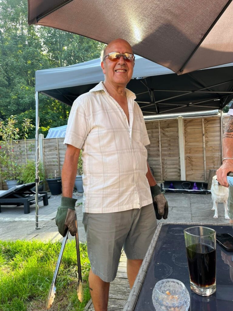 A man in a white shirt (Charlie) smiles at the camera. Charlie is in the garden and is stood in front of a patio table, he is wearing oven gloves and holding a cooking utensil.
