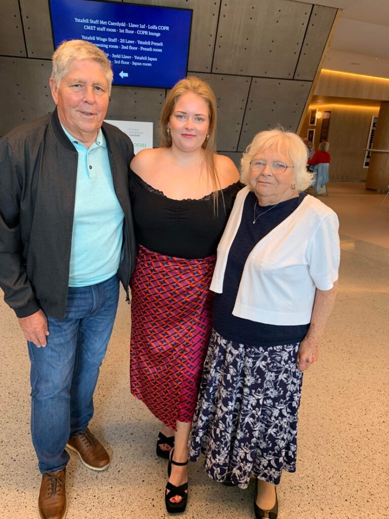Charlotte is pictured with her grandparents. Charlotte stands in the middle and wears a red, patterned skirt and black, off the shoulder top, with black heeled sandals. She has long blonde hair and smiles at the camera. Her grandad is wearing jeans, brown smart shoes, a blue t-shirt and a black, long-sleeved jacket. Her grandma is wearing a navy, floral skirt, with a black plain top and white, short sleeve cardigan, with black shoes.