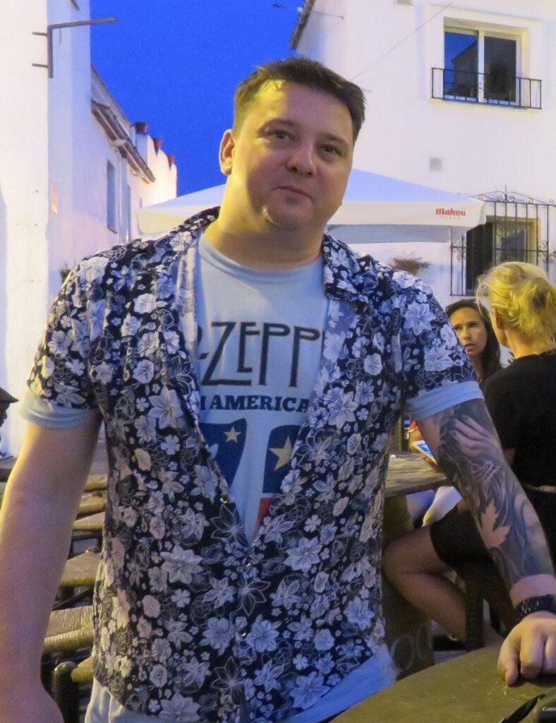 Mark is outside at dusk at wears a white Led Zeppelin t-shirt, with a black and white floral short-sleeved shirt on top, partially buttoned up. He has a sleeve of tattoos on his left arm and short, light brown hair which is spiked up. He smiles slightly at the camera.