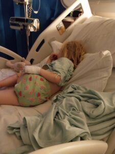 Mia is lying on her side in a hospital bed, wearing a mint-green top and watermelon-patterned shorts. Her left hand is wrapped in a white medical dressing.