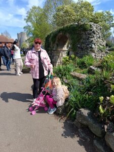 Larissa stands beside Mia, who is sitting on some stones. Larissa is wearing sunglasses, a pink jacket, and black trousers. Mia is wearing a pink floral jacket and pink trousers, with a pink toy stroller next to her. They are outdoors, surrounded by greenery, with a stone structure in the background.