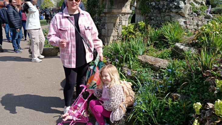 Larissa stands beside Mia, who is sitting on some stones. Larissa is wearing sunglasses, a pink jacket, and black trousers. Mia is wearing a pink floral jacket and pink trousers, with a pink toy stroller next to her. They are outdoors, surrounded by greenery, with a stone structure in the background.