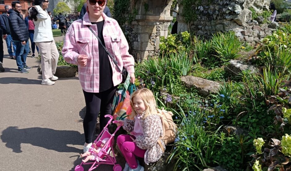 Larissa stands beside Mia, who is sitting on some stones. Larissa is wearing sunglasses, a pink jacket, and black trousers. Mia is wearing a pink floral jacket and pink trousers, with a pink toy stroller next to her. They are outdoors, surrounded by greenery, with a stone structure in the background.