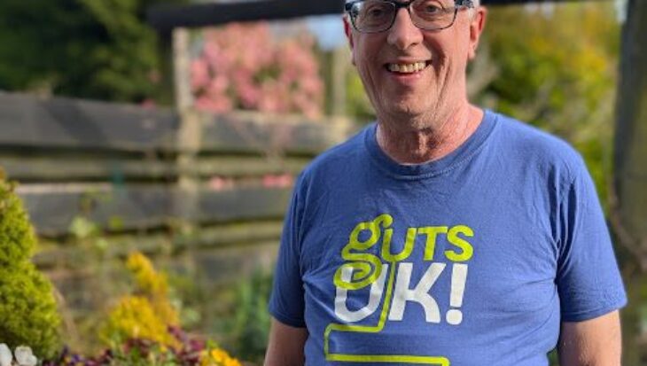 Richard is stood in is garden, wearing a blue Guts UK t-shirt, grey trousers and black rimmed glasses. He has grey hair, and a silver watch on his arm. He has his hands in his pockets and to his left, are a selection of purple flowers, and other shrubs in pots, on a garden table. He smiles at the camera.