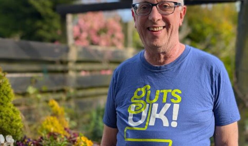 Richard is stood in is garden, wearing a blue Guts UK t-shirt, grey trousers and black rimmed glasses. He has grey hair, and a silver watch on his arm. He has his hands in his pockets and to his left, are a selection of purple flowers, and other shrubs in pots, on a garden table. He smiles at the camera.