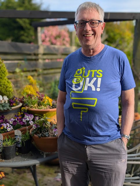 Richard is stood in is garden, wearing a blue Guts UK t-shirt, grey trousers and black rimmed glasses. He has grey hair, and a silver watch on his arm. He has his hands in his pockets and to his left, are a selection of purple flowers, and other shrubs in pots, on a garden table. He smiles at the camera.