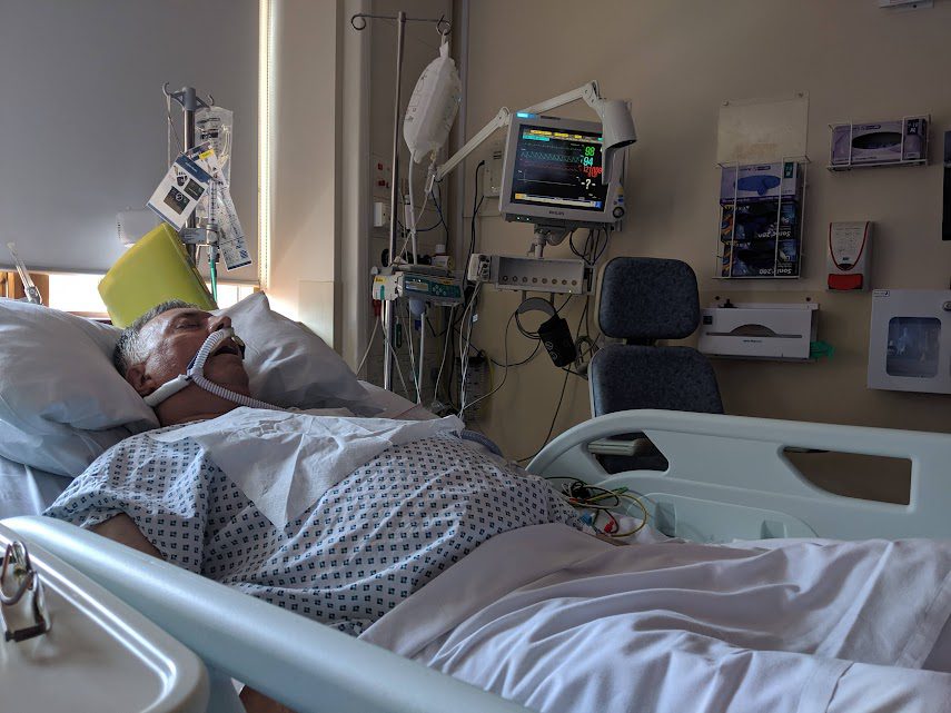 Richard is in a hospital bed in the ITU. He has monitors around him, and is asleep, with oxygen going into his nose. He wears a white hospital gown with small teal diamonds on, and his bed is in front of a window with a blind that is mostly closed. There is also a drip trolley with medication on that Richard is receiving via a line into his body.