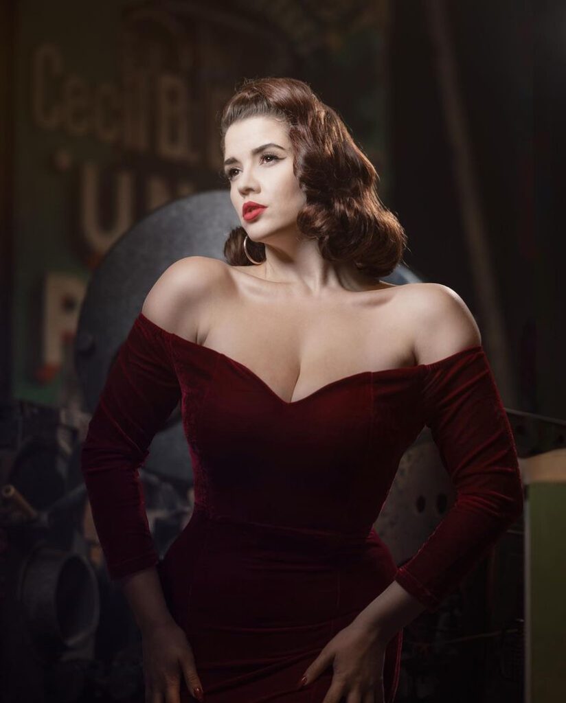 Scarlett poses against a dark background. She has a vintage, off the shoulder, dark red velvet dress on. Her dark brown hair is curled loosely, and fixed into place by the sides of her face. She is looking towards the left of the shot, pouting with red lips and she also has gold hoop earrings in.