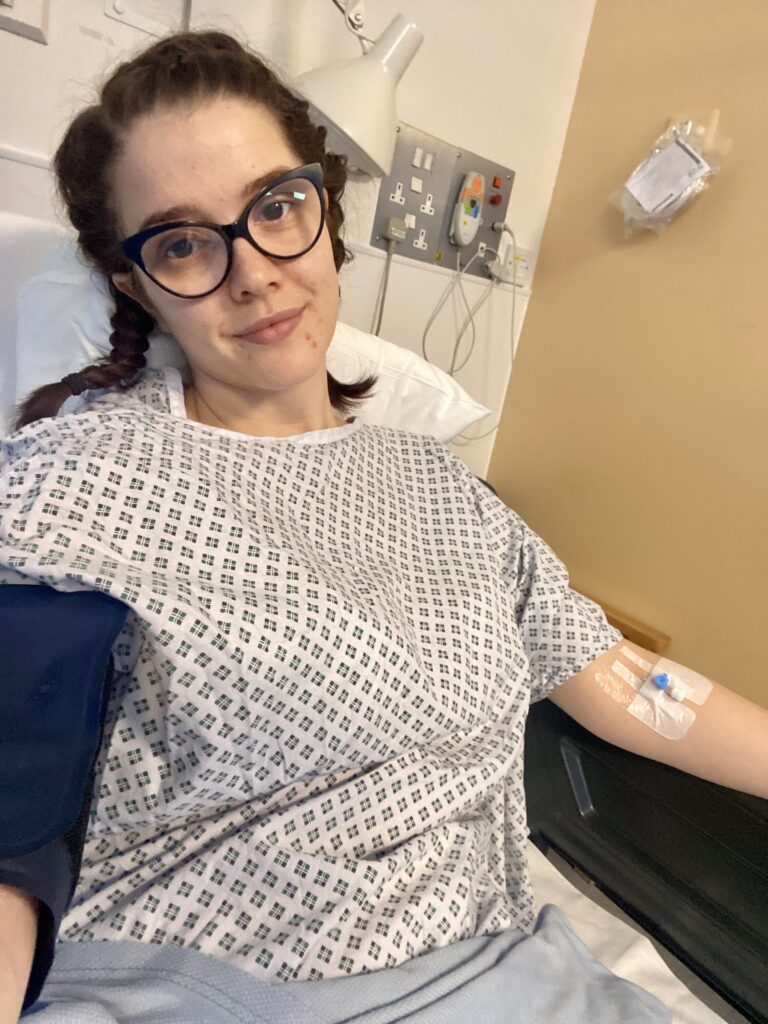 Scarlett sits partially upright on a hospital bed. She wears a white hospital gown with blue diamonds on, a dark blue blood pressure sleeve and she has a cannula in her opposite arm. She has dark brown hair, in plaits, black rimmed oval glasses and smiles at the camera.