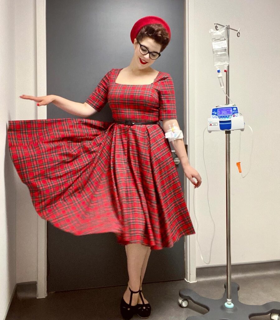 Scarlett is in hospital, connected to an intravenous drip going into her arm, to receive her Crohn's disease treatment. Her drip stand is beside her. She is stood smiling towards the floor, in a three quarter sleeve vintage, red tartan dress and has red nails to match. A thin black belt cinches her dress in at the waist, and she also wears a bright red beret style hat and black patent Mary Jane style heels. She wears red lipstick and thick, black rimmed oval glasses. One arm is down by her side and the other has just let one side of her tartan dress down from her waist, to reveal the full skirt.