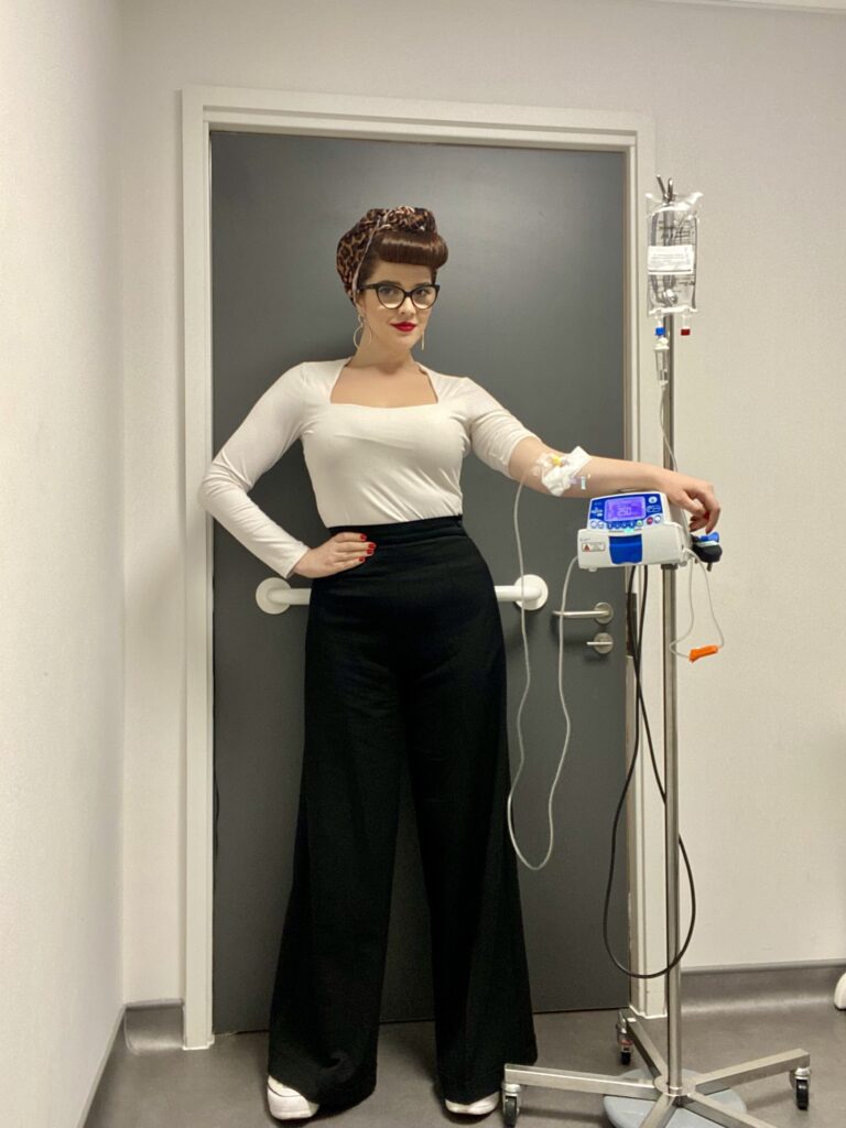 Scarlett is in hospital, connected to an intravenous drip for her Crohn's treatment. She wears a white long sleeved top, black, wide-leg, high-waisted trousers and white trainers. She has red nails, and has her dark brown hair up, held by a leopard print bandana, with her fringe curled over her forehead. She also wears gold hoop earrings, red lipstick and black rimmed oval glasses. She has one hand on her hop, posing proudly, and her other arm resting on top of her treatment machine on her drip stand.