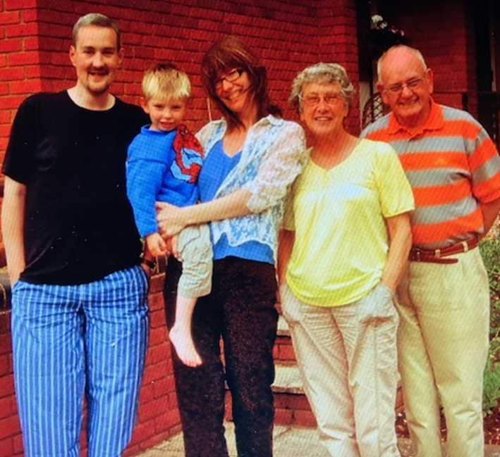 group of five people, appearing to be a multi-generational family, standing outdoors against a red brick wall. On the left is a tall man in a black shirt and blue striped trousers. Next to him, a woman with glasses holds a small boy in a blue and red top. To the right are an older woman in a yellow top and an older man in an orange and grey striped shirt.