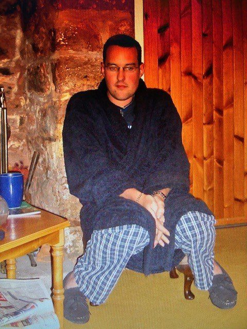 A man, Andrew, wearing a dark dressing gown and plaid pajama trousers, sitting near a fire pit or stone wall. He is wearing glasses and has his hands clasped on his knees. A wooden wall is visible to the right.