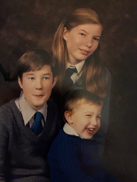 A portrait of three children. A girl with long hair and a school uniform is in the background. In the foreground, a boy in a grey sweater and tie is on the left, and a younger boy with a wide smile in a blue top is on the right.