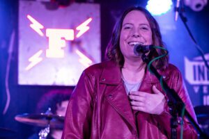 Beth is wearing a burgundy leather jacket and passionately singing into a microphone on stage. She has her left hand on her chest and is standing in front of a sign that says "F" with her bandmate in the background. 