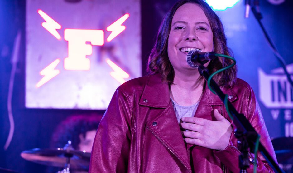 Beth is wearing a burgundy leather jacket and passionately singing into a microphone on stage. She has her left hand on her chest and is standing in front of a sign that says "F" with her bandmate in the background.