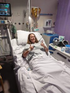 Beth is laying down in a hospital bed, giving a thumbs up and smiling at the camera. She is hooked to some medical equipment and has a tube in her nose. 