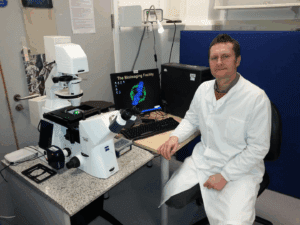 Dr Jason Bruce sits at a desk wearing a white lab coat. He is sat next to science equipment and a computer.