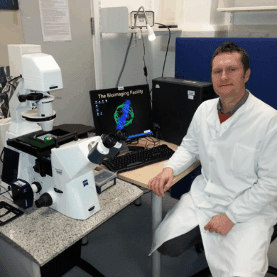 Dr Jason Bruce sits at a desk wearing a white lab coat. He is sat next to science equipment and a computer.