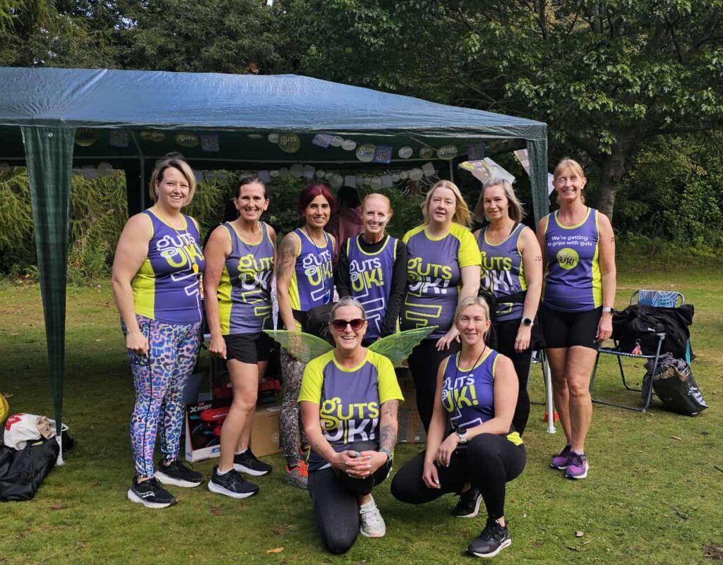 A group of nine women, including Eryn, wearing matching blue and green "Guts yellow!" t-shirts and vests pose for a photo outdoors. Most are standing, while two women are kneeling in the front. They are standing on grass under a green canopy tent decorated with Guts UK bunting. To the left, there's a table with wrapped raffle gifts. To the right, a white 4x4 car with its boot open is visible. Trees and greenery are in the background.