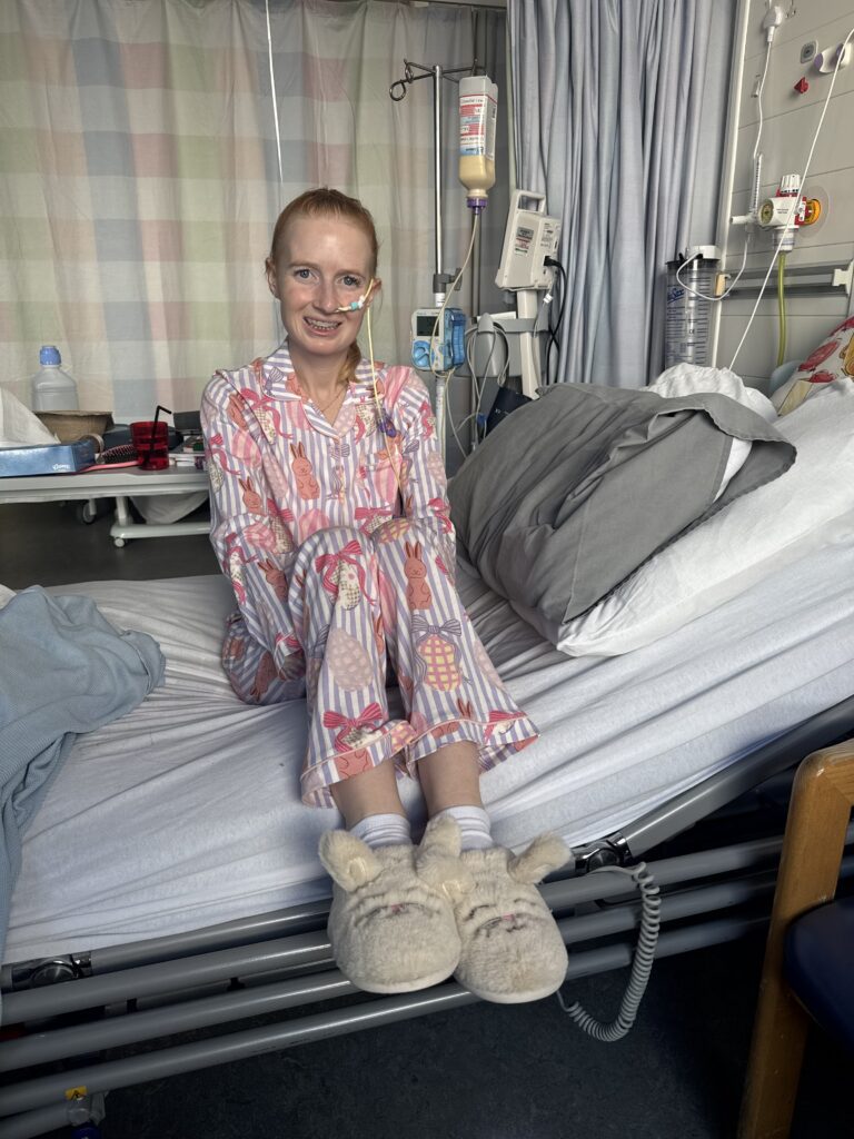 Eryn is smiling. She has reddish hair sits on a hospital bed, facing forward. She is wearing pink pyjamas with bunny and Easter egg patterns, and fluffy white bunny slippers. A feeding tube is visible, going into Eryn's nostril, and an IV drip stand with medical equipment is in the background. The bed has white sheets and a grey pillow.