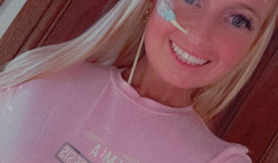 Eryn is smiling and has long blonde hair, wearing a pink t-shirt with text. She has a feeding tube visible on her face, taped near her nose and cheek, and long earrings. The text on her shirt reads 