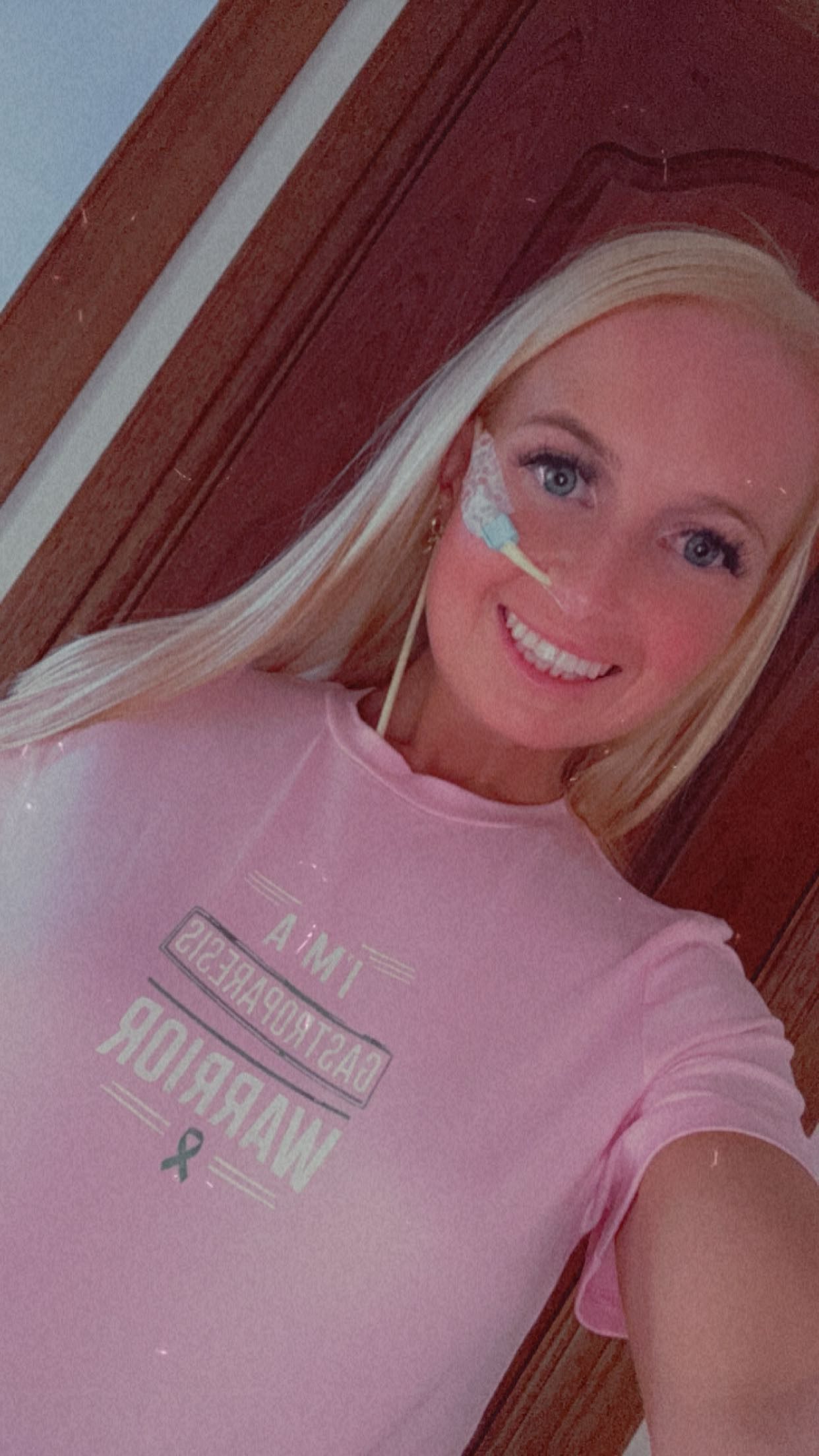 Eryn is smiling and has long blonde hair, wearing a pink t-shirt with text. She has a feeding tube visible on her face, taped near her nose and cheek, and long earrings. The text on her shirt reads "I'm a gastroparesis warrior" with a ribbon symbol. The background is a warm-toned wooden door or wall.