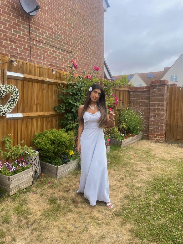 Esha stands in a garden wearing a long, elegant light blue dress and white sandals. She has long brown hair with a white flower clip in it and Esha also wears a necklace. Behind her, there is a wooden fence decorated with a heart-shaped wreath and blooming pink roses, with houses visible in the background under a cloudy sky.