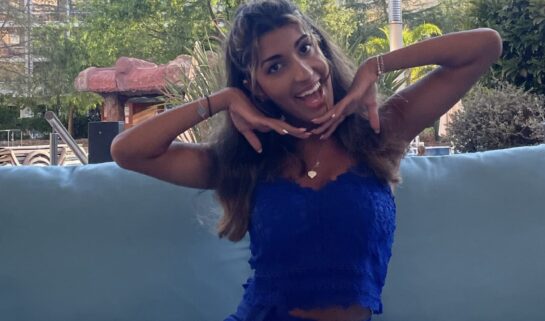 Esha is sat on a light blue sofa, outdoors, abroad. The sun is shining and she wears an electric blue two piece outfit, consisting of a vest and a short skirt. She has long, wavy brunette hair and holds her hands under her chin, smiling widely at the camera.