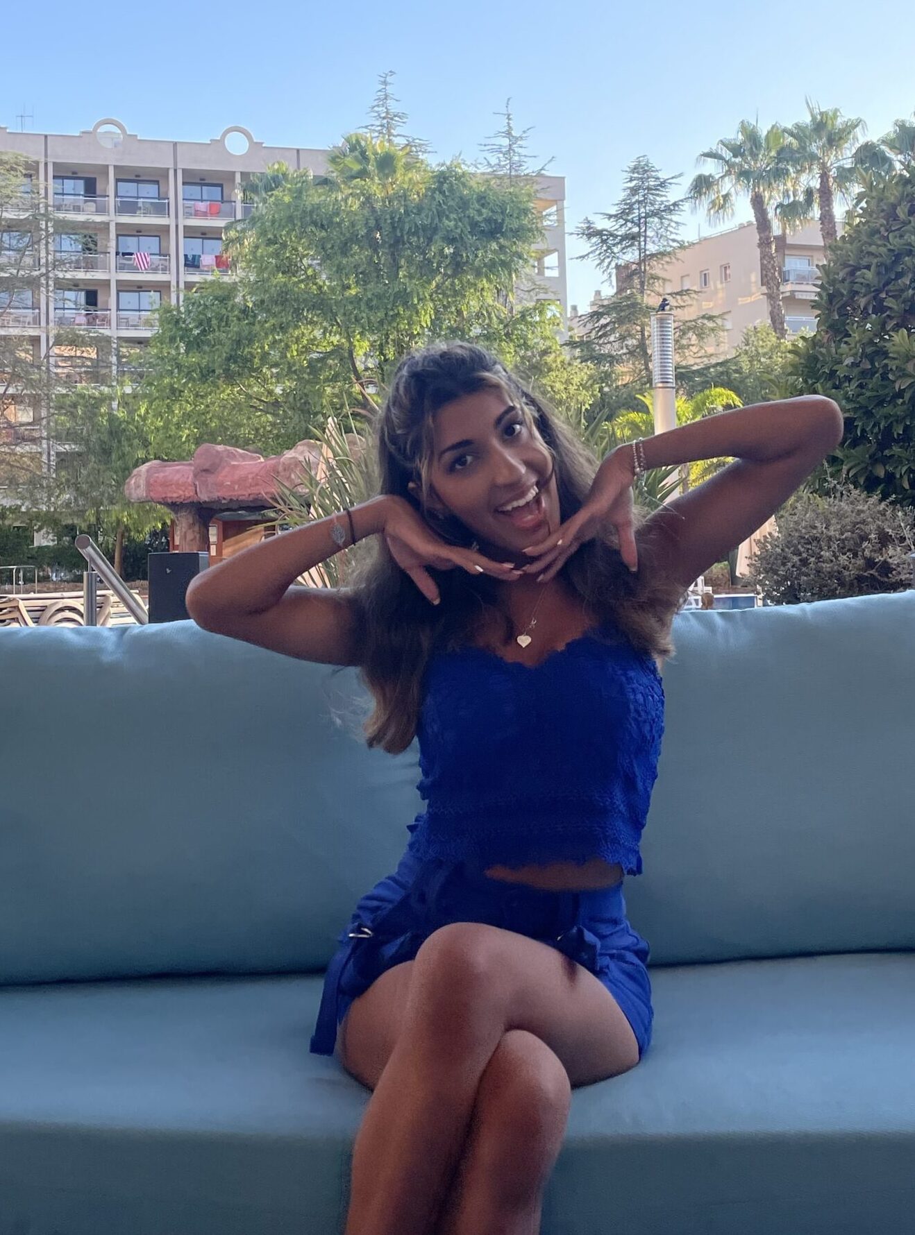 Esha is sat on a light blue sofa, outdoors, abroad. The sun is shining and she wears an electric blue two piece outfit, consisting of a vest and a short skirt. She has long, wavy brunette hair and holds her hands under her chin, smiling widely at the camera.