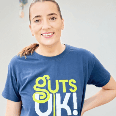 Scarlett stands in front of a white wall, wearing a navy Guts UK t-shirt. She smiles and has one hand on her hip. She has brown, long hair, scooped back off her face in a ponytail.