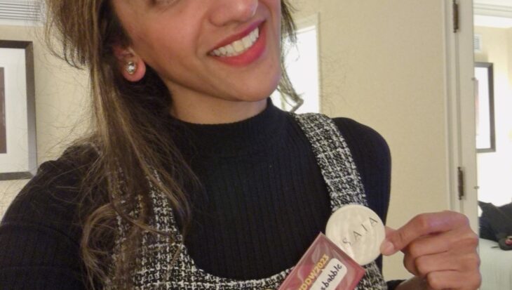 Sharan, smiling directly at the camera in a room, wearing a black turtleneck under a black and white checked pinafore dress. Attached to her dress are two badges, one of which reads 