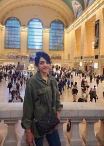 Sharan is smiling and wearing an oversized green corduroy jacket and jeans, with a quilted black shoulder bag, standing inside Grand Central Terminal. The large, arched windows and vaulted ceiling of the terminal are visible in the background, filled with people.