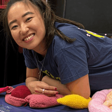 Leeona has long dark hair tied up with a red scrunchie and smiles directly at the camera. She is leaning over a table covered with a dark blue cloth, resting her arms on it. She is wearing a navy blue t-shirt and has some knitted guts and Guts UK leaflets in front of her. In the background, part of a monitor screen is visible.