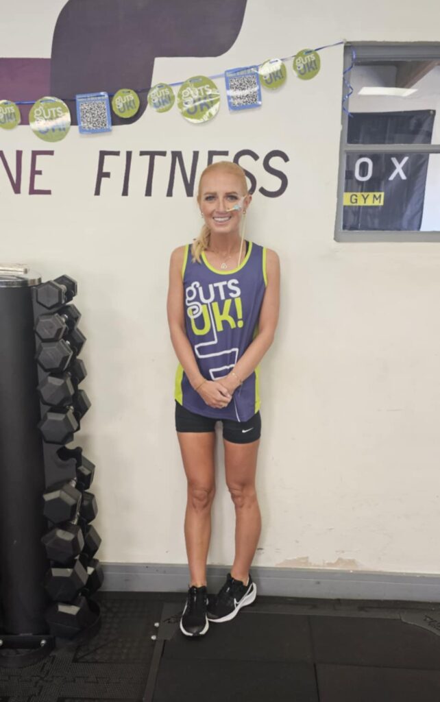 Eryn is standing in a gym wearing a blue Guts UK running vest and black shorts. She also wears black and white trianers.